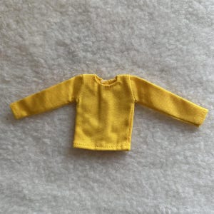 Puede incluir: Una camisa amarilla brillante de manga larga. La camisa est&aacute; hecha de una tela suave y tiene cuello redondo. Las mangas son largas y llegan hasta las mu&ntilde;ecas. La camisa est&aacute; dise&ntilde;ada para una mu&ntilde;eca.