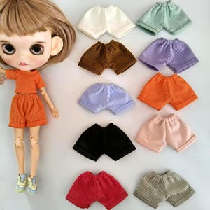 May include: A doll with brown hair wearing an orange ribbed t-shirt and matching shorts, displayed alongside ten additional pairs of ribbed shorts in various solid colors including white, green, brown, purple, orange, light blue, black, pink, red, and gray.