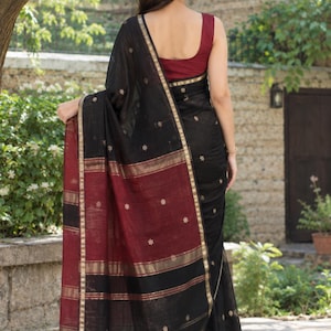 May include: A black saree featuring a deep red border with gold metallic stripes and small embroidered floral motifs. The garment includes a sleeveless maroon blouse and a draped black fabric panel with black tassels at the bottom edge.