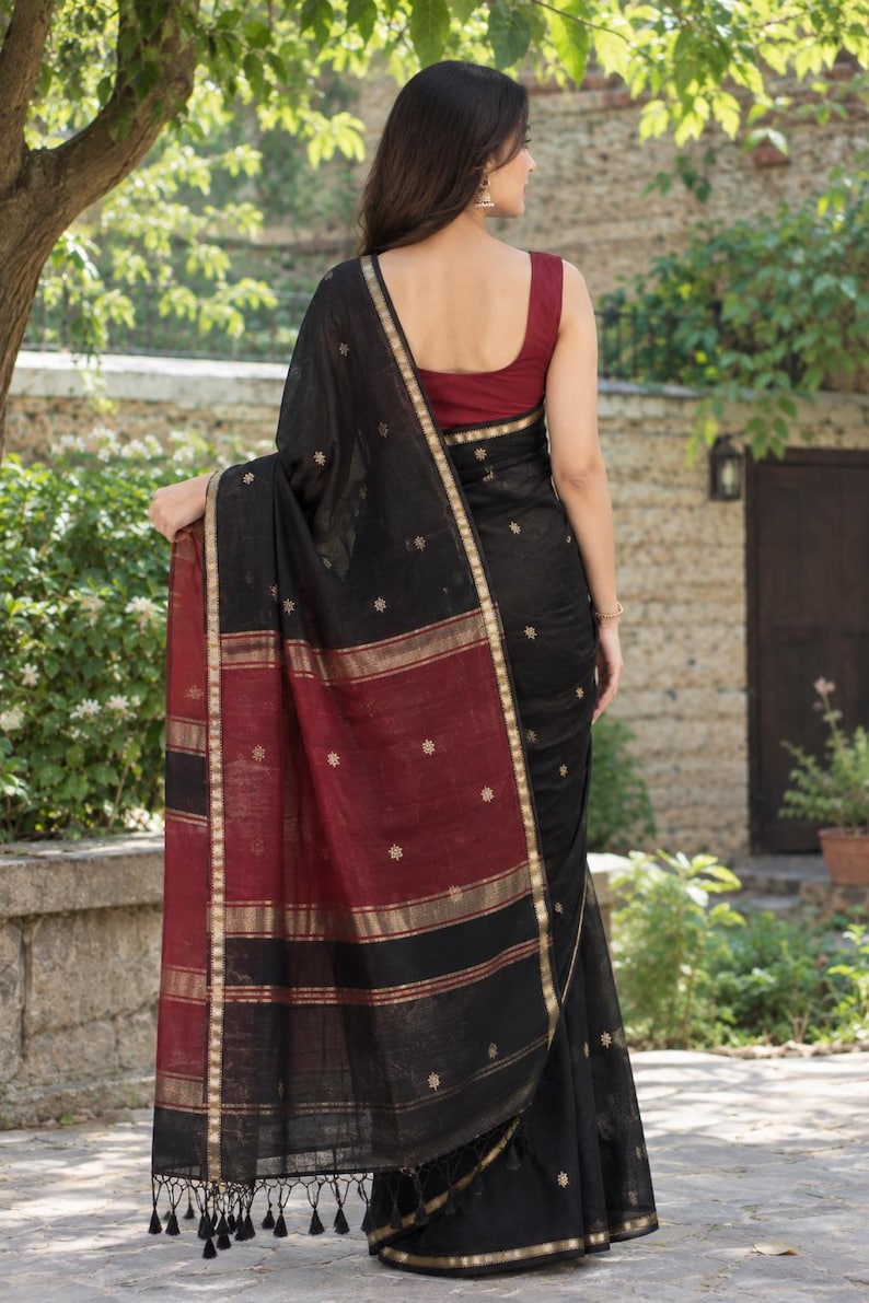 May include: A black saree featuring a deep red border with gold metallic stripes and small embroidered floral motifs. The garment includes a sleeveless maroon blouse and a draped black fabric panel with black tassels at the bottom edge.