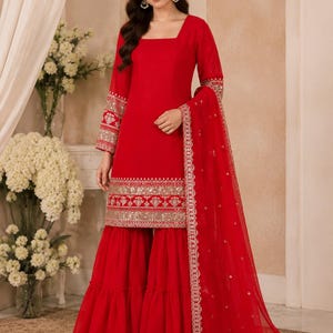 May include: A vibrant red long-sleeved tunic featuring a square neckline and intricate gold embroidery along the cuffs and hem. The outfit includes matching wide-leg flared trousers and a sheer red dupatta with a decorative gold embroidered border.