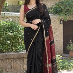May include: A black saree featuring small gold embroidered floral motifs, a gold border, and a contrasting maroon pallu with black and gold horizontal stripes. The garment includes black tassels at the end of the pallu and is worn with a maroon blouse.