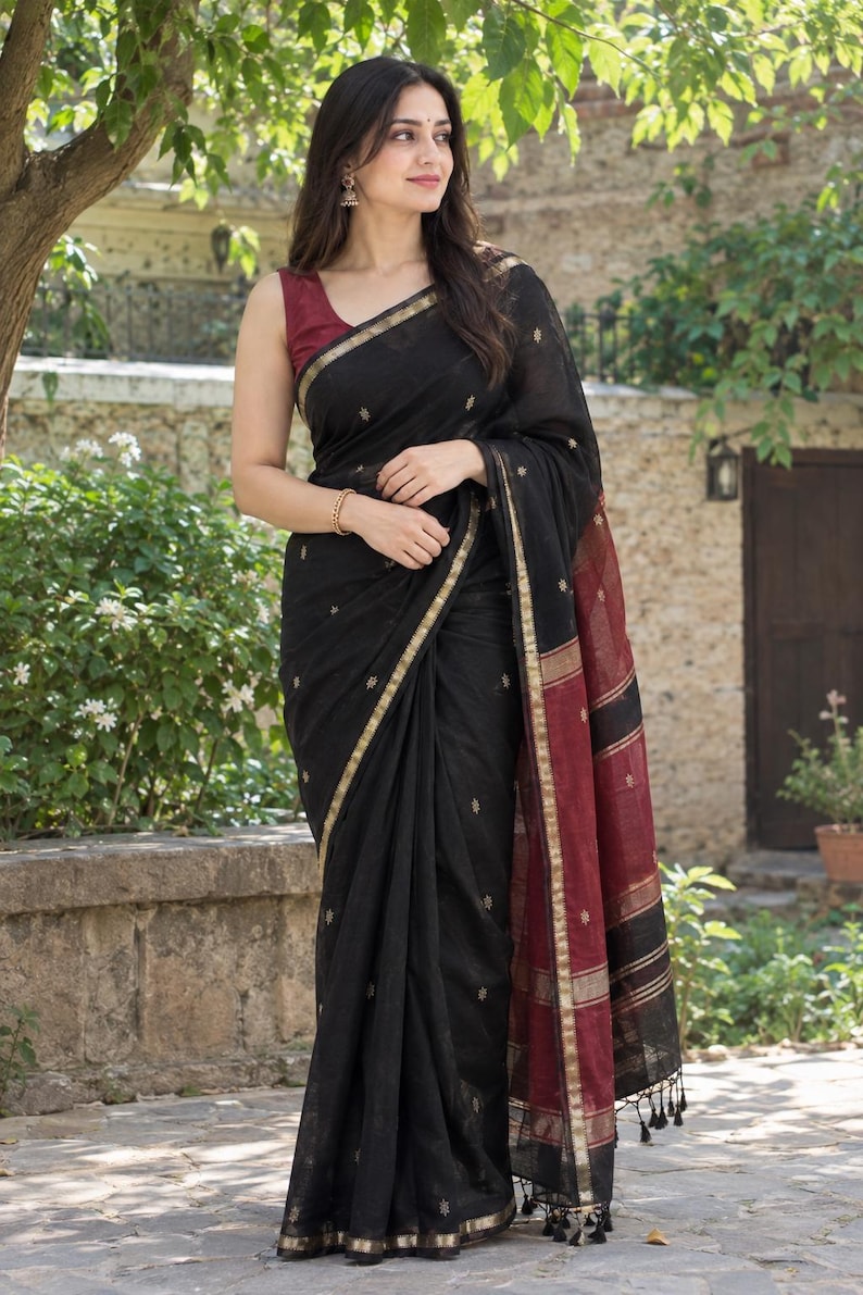 May include: A black saree featuring small gold embroidered floral motifs, a gold border, and a contrasting maroon pallu with black and gold horizontal stripes. The garment includes black tassels at the end of the pallu and is worn with a maroon blouse.
