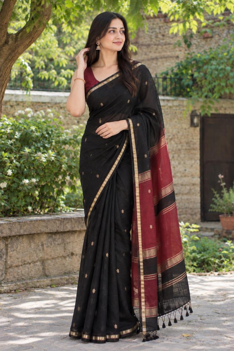 May include: A black saree featuring small gold embroidered motifs throughout the fabric, a gold border, and a wide maroon end piece with horizontal gold stripes and black tassels at the hem, paired with a sleeveless maroon blouse.