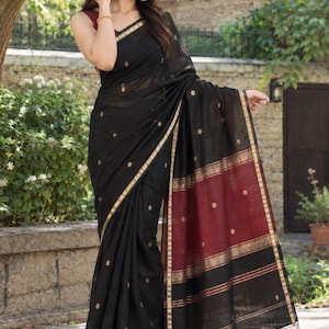 May include: A black saree featuring small gold floral motifs, a wide maroon border with gold stripes, and a gold-trimmed edge. The garment includes a maroon blouse and a hem finished with black tassels, showcasing a traditional drape and texture.