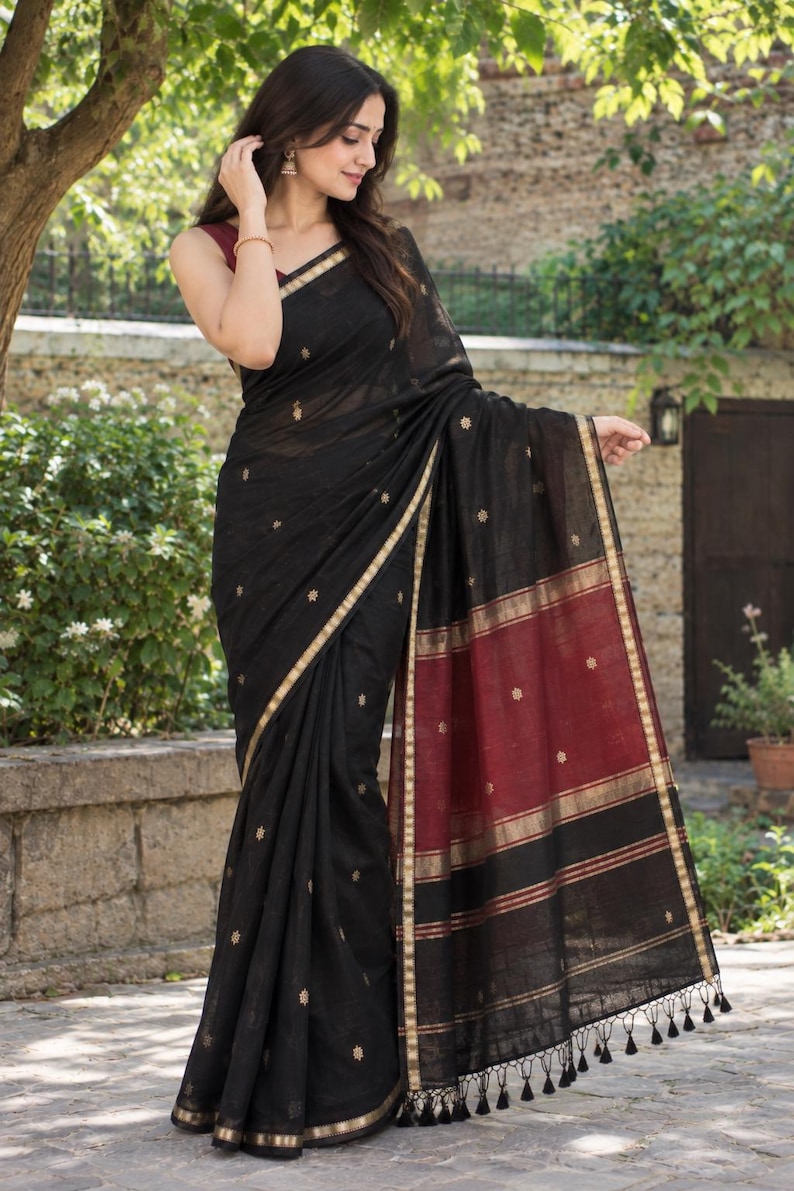 May include: A black saree featuring small gold floral motifs, a wide maroon border with gold stripes, and a gold-trimmed edge. The garment includes a maroon blouse and a hem finished with black tassels, showcasing a traditional drape and texture.