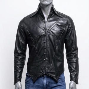 May include: A black leather jacket with a unique button-up design. The jacket features a pointed collar and long sleeves. The jacket is displayed on a mannequin wearing blue jeans. The jacket has a distinctive, tailored fit.