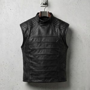 Op de afbeelding: Een zwart leren vest met een hoge kraag en een mouwloos ontwerp. Het vest heeft horizontale en geometrische stikseldetails, hangend aan een metalen hanger tegen een grijze muur.