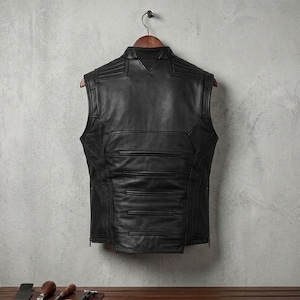 Op de afbeelding: Een zwart leren vest met een op maat gemaakt ontwerp hangt aan een houten hanger. Het vest heeft een hoge kraag, horizontale stiksels en een ritssluiting. Het vest is mouwloos en lijkt nieuw.