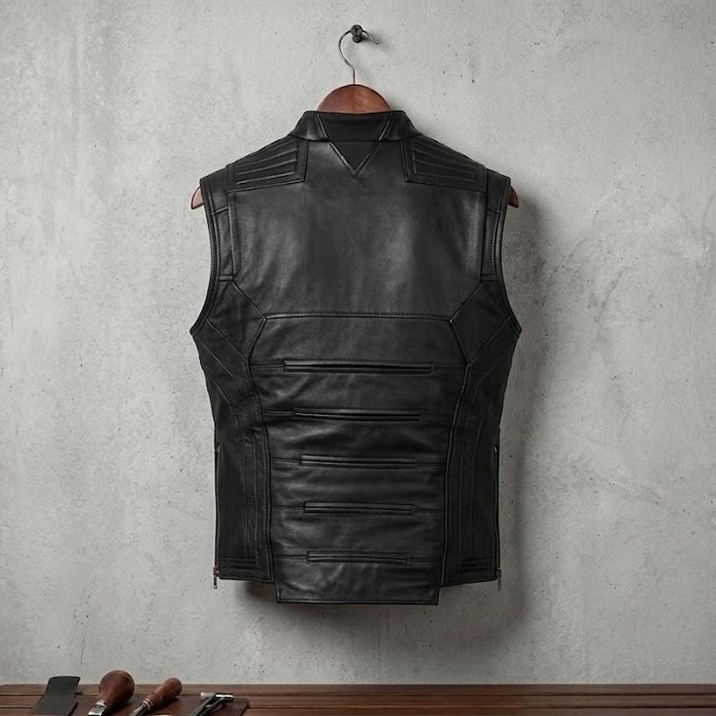 Op de afbeelding: Een zwart leren vest met een op maat gemaakt ontwerp hangt aan een houten hanger. Het vest heeft een hoge kraag, horizontale stiksels en een ritssluiting. Het vest is mouwloos en lijkt nieuw.