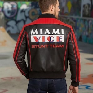 May include: Black leather jacket with red accents and the text "MIAMI VICE STUNT TEAM" on the back. The jacket has a ribbed design on the shoulders and arms, and a black collar and cuffs. The model is wearing blue jeans.