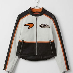 May include: A white, black, and orange racing jacket with a full-length zipper. The jacket features the McLaren logo on the chest and orange stripes down the sleeves. The collar and interior lining are orange.