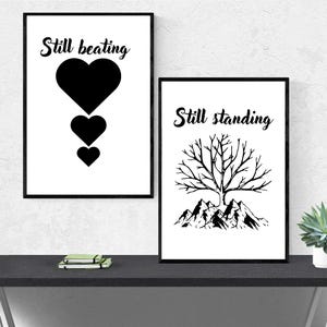 May include: Two framed wall art prints with black and white designs. The left print features the text Still beating above three stacked black hearts. The right print displays the text Still standing above a black tree silhouette over mountains.