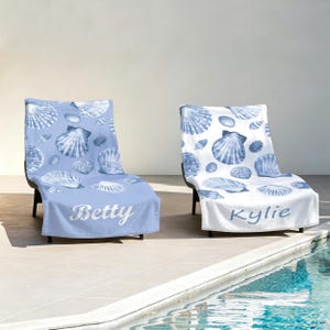 Peut inclure: Deux serviettes de plage drapées sur des chaises longues. Une serviette est bleu clair avec des motifs de coquillages blancs et le prénom Betty en écriture blanche. L'autre est blanche avec des motifs de coquillages bleus et le prénom Kylie en bleu.