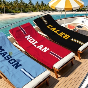 Può includere: Tre teli mare rettangolari in azzurro, rosso e nero, ognuno con un nome in bianco o giallo stampato in un carattere atletico in grassetto. I teli sono decorati con due strisce parallele e sono appoggiati su sedie a sdraio in legno su una barca.