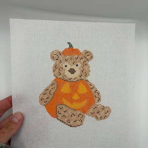 Puede incluir: Un lienzo de punto de aguja que muestra un oso de peluche color canela con un disfraz de calabaza naranja. El oso tiene una nariz marrón y ojos oscuros, mientras que el traje de calabaza presenta una cara de linterna con ojos y boca amarillos.