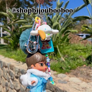 May include: A handmade keychain featuring a Bratz doll in a blue outfit with a white fur trim, a blue pom-pom, and a Cinnamoroll charm. The keychain includes colorful beads and the text "Bijou Boo Boo".