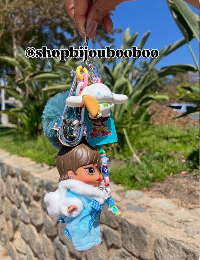 May include: A handmade keychain featuring a Bratz doll in a blue outfit with a white fur trim, a blue pom-pom, and a Cinnamoroll charm. The keychain includes colorful beads and the text "Bijou Boo Boo".