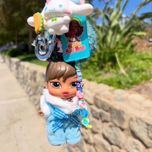 May include: A keychain featuring a Bratz doll in a blue outfit, a white cartoon character, a taco charm, and a blue pom-pom. The keychain also includes a charm with the text "Bijou Boo Boo". The bottom of the image has the text "@shopbijoubooboo".