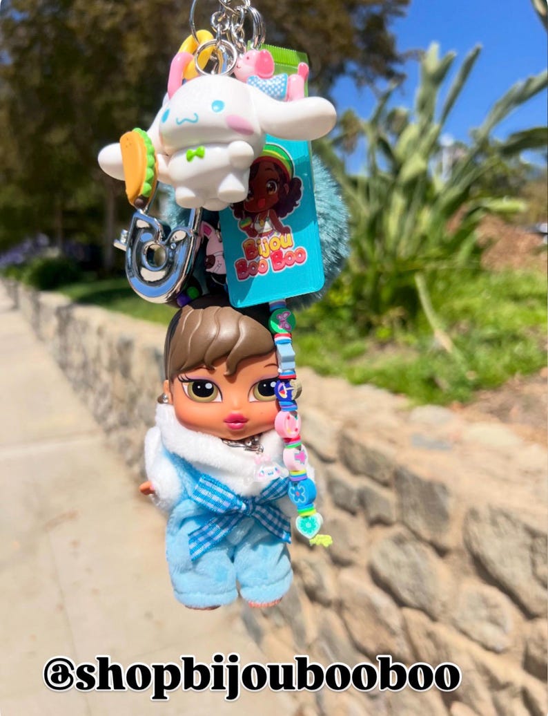 May include: A keychain featuring a Bratz doll in a blue outfit, a white cartoon character, a taco charm, and a blue pom-pom. The keychain also includes a charm with the text "Bijou Boo Boo". The bottom of the image has the text "@shopbijoubooboo".