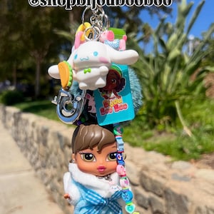 May include: A multi-charm keychain featuring a Bratz doll, a white cartoon character, and other colorful charms. The Bratz doll wears a blue outfit with a white fur collar. The keychain includes a silver ring and the text "@shopbijoubooboo".