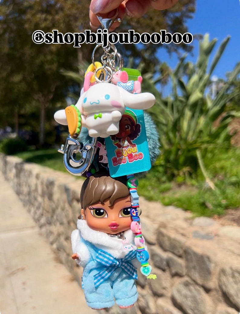 May include: A multi-charm keychain featuring a Bratz doll, a white cartoon character, and other colorful charms. The Bratz doll wears a blue outfit with a white fur collar. The keychain includes a silver ring and the text "@shopbijoubooboo".