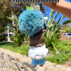 May include: A keychain featuring a teal pom-pom, a brown-haired doll, and a plush figure with blue plaid pants. The keychain is held up in front of a blurred outdoor background. The text "@shopbijoubooboo" is at the top.