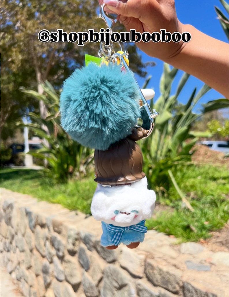 May include: A keychain featuring a teal pom-pom, a brown-haired doll, and a plush figure with blue plaid pants. The keychain is held up in front of a blurred outdoor background. The text "@shopbijoubooboo" is at the top.