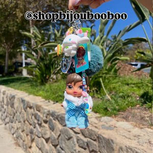 May include: A keychain featuring a Bratz doll in blue overalls and a white top, along with a white cartoon character, a teal pom-pom, and other charms. The text "@shopbijoubooboo" is visible.