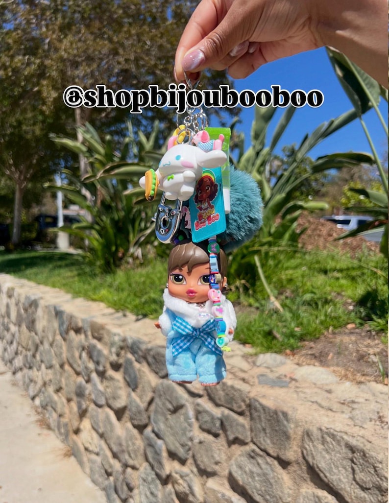 May include: A keychain featuring a Bratz doll in blue overalls and a white top, along with a white cartoon character, a teal pom-pom, and other charms. The text "@shopbijoubooboo" is visible.