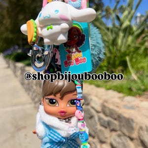May include: A keychain featuring a Bratz doll, a white cartoon character with a taco, a pink pig, and a blue pom-pom. The keychain has a silver clasp and the text "@shopbijoubooboo".