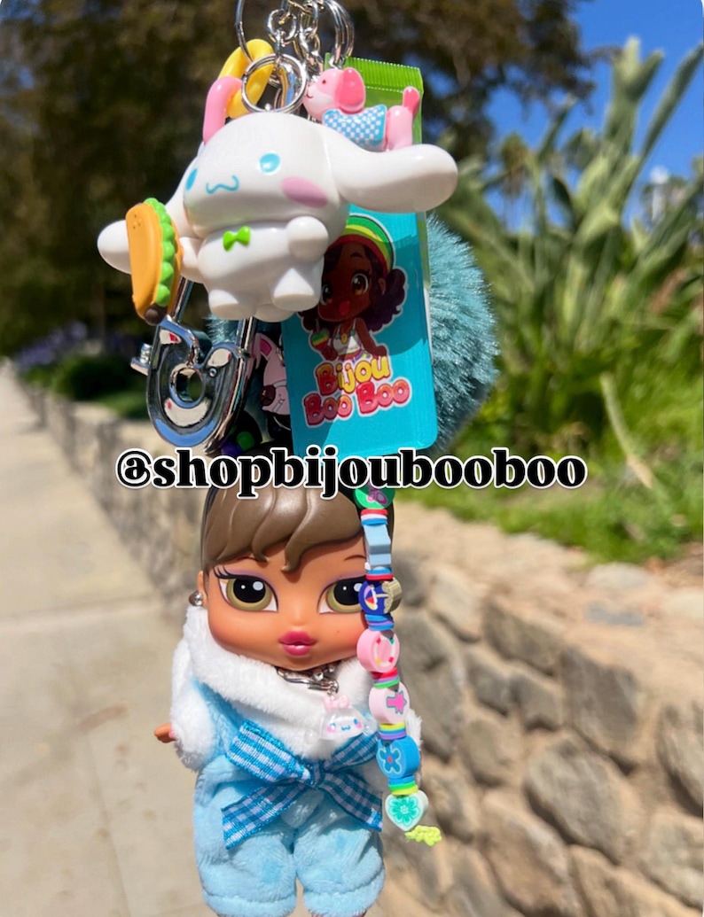 May include: A keychain featuring a Bratz doll, a white cartoon character with a taco, a pink pig, and a blue pom-pom. The keychain has a silver clasp and the text "@shopbijoubooboo".