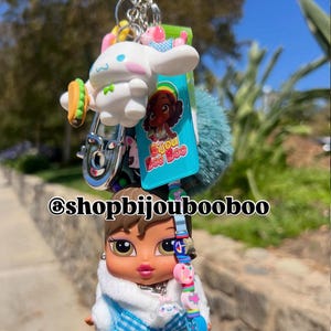 May include: A keychain featuring a Bratz doll wearing a blue outfit with a white fur-trimmed jacket. The keychain also includes a white cartoon character with a taco, a blue pom-pom, and a charm with the text "Bijou Boo Boo".