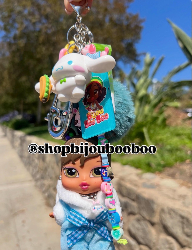 May include: A keychain featuring a Bratz doll wearing a blue outfit with a white fur-trimmed jacket. The keychain also includes a white cartoon character with a taco, a blue pom-pom, and a charm with the text "Bijou Boo Boo".