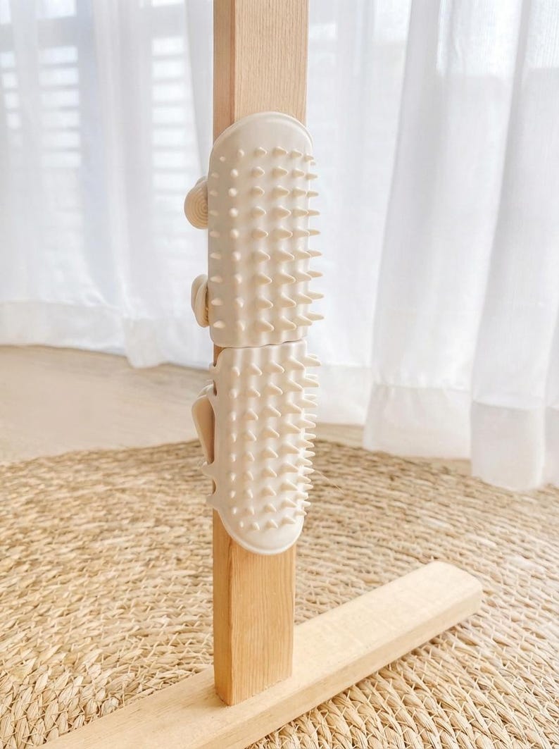 May include: A beige, spiky, two-piece furniture protector attached to a light wood post. The protector has a series of rounded spikes and is secured with small, curved clasps. The background includes a woven mat and white curtains.