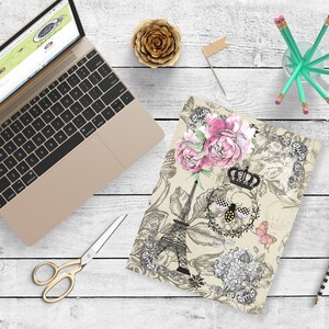 May include: A beige notebook cover with a black and white illustration of the Eiffel Tower, flowers, and a bee with a crown. The design has pink and black accents.