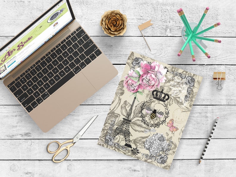 May include: A beige notebook cover with a black and white illustration of the Eiffel Tower, flowers, and a bee with a crown. The design has pink and black accents.