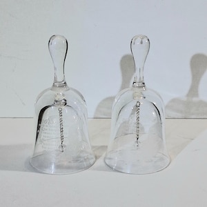 May include: Two clear glass bell-shaped ornaments with long, curved handles. Each bell has a beaded clapper and etched text. The bells are displayed on a white surface. The text on the bell reads "Be kind..."