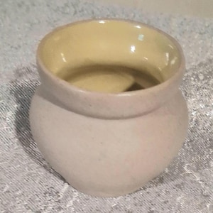 May include: A small, round, ceramic pot with a light beige exterior and a pale yellow interior. The pot has a slightly textured surface and a wide opening. The design is simple and functional, suitable for various uses.