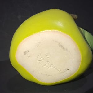 May include: A ceramic pear-shaped decorative item. The pear is a bright yellow-green color with a white base. A small green leaf and stem are attached. The bottom of the pear has the inscription "G 2000".