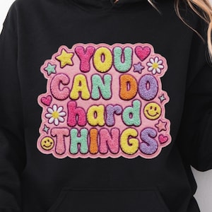 May include: Black hoodie featuring the text "YOU CAN DO HARD THINGS" in a colourful graphic. The design includes stars, hearts, flowers, and a smiley face. The hoodie has a front pocket and a hood.