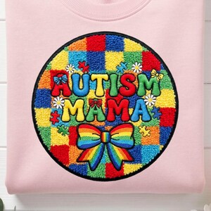 Peut inclure: Un sweat-shirt rose clair avec un motif circulaire. Le motif présente les mots "AUTISM MAMA" en couleurs, un nœud arc-en-ciel et des pièces de puzzle. Le fond est composé de carrés colorés.