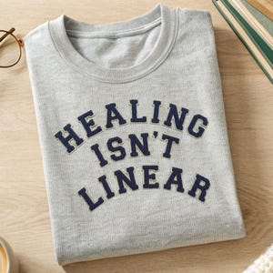 Puede incluir: Una sudadera gris claro doblada con el texto azul marino "HEALING ISN'T LINEAR". La sudadera está sobre una superficie de madera clara, con un par de gafas y un libro en el fondo.
