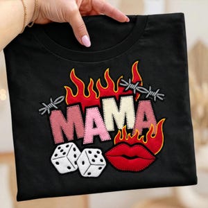May include: Black t-shirt with an embroidered design featuring the word "MAMA" in pink and white, surrounded by flames and barbed wire. Includes dice and red lips.