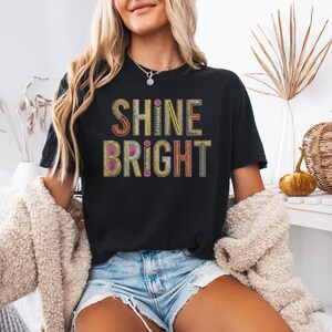 May include: Black t-shirt with the words "SHINE BRIGHT" in colorful, textured lettering. The shirt is worn by a person, paired with denim shorts and a cream-colored cardigan. The background includes decorative elements.