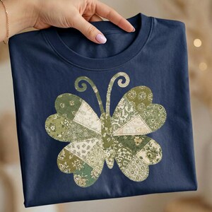 May include: A navy blue t-shirt with a butterfly motif. The butterfly is made from green and cream-coloured fabric patches with floral and patterned designs. The shirt has a crew neck and short sleeves.