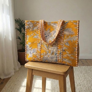 May include: A large, quilted tote bag with a vibrant yellow background and a floral pattern in shades of blue and white. The bag has sturdy handles with a striped design and is sitting on a wooden stool.