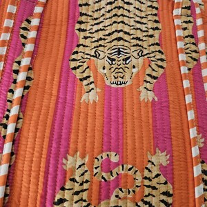 May include: Quilted fabric with a tiger print. The design features a large tiger in the center, with smaller tiger motifs at the bottom. The background has vertical stripes in orange and pink, with white and orange striped borders.