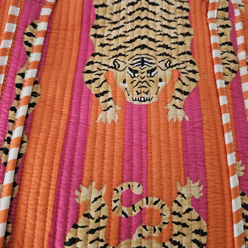 May include: Quilted fabric with a tiger print. The design features a large tiger in the center, with smaller tiger motifs at the bottom. The background has vertical stripes in orange and pink, with white and orange striped borders.