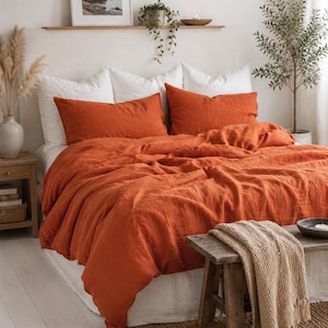 May include: A bed featuring a vibrant burnt orange duvet cover and two matching rectangular pillow shams. The bedding is layered over white pillows and a white bed skirt, creating a textured appearance on the fabric surface of the linens.